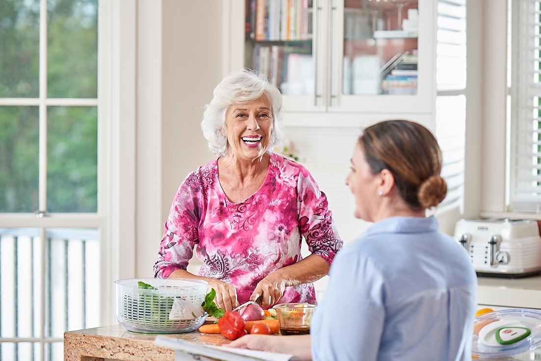 An elderly woman smiling while preparing food in a bright kitchen, engaging with a female home care worker.